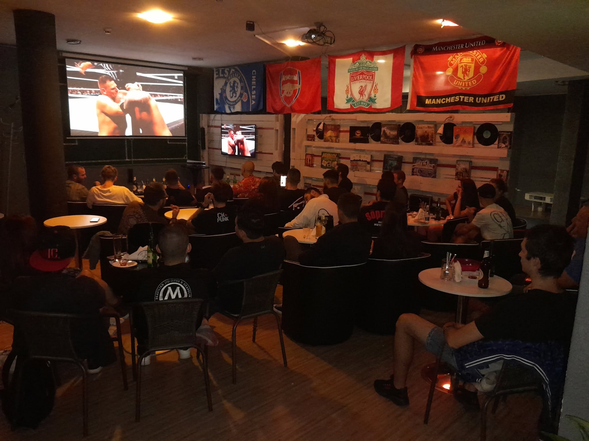 Пълен салон гледа UFC мач на проектора под знамената на английските клубове.